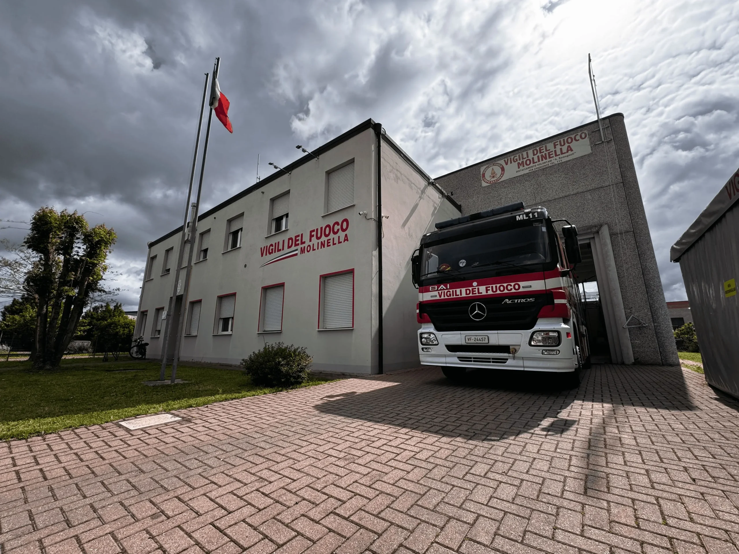 Camion dei vigili del fuoco fermo nel parcheggio davanti alla caserma, con una porzione visibile dell'entrata, fotografato nel primo pomeriggio di un giorno annuvolato.